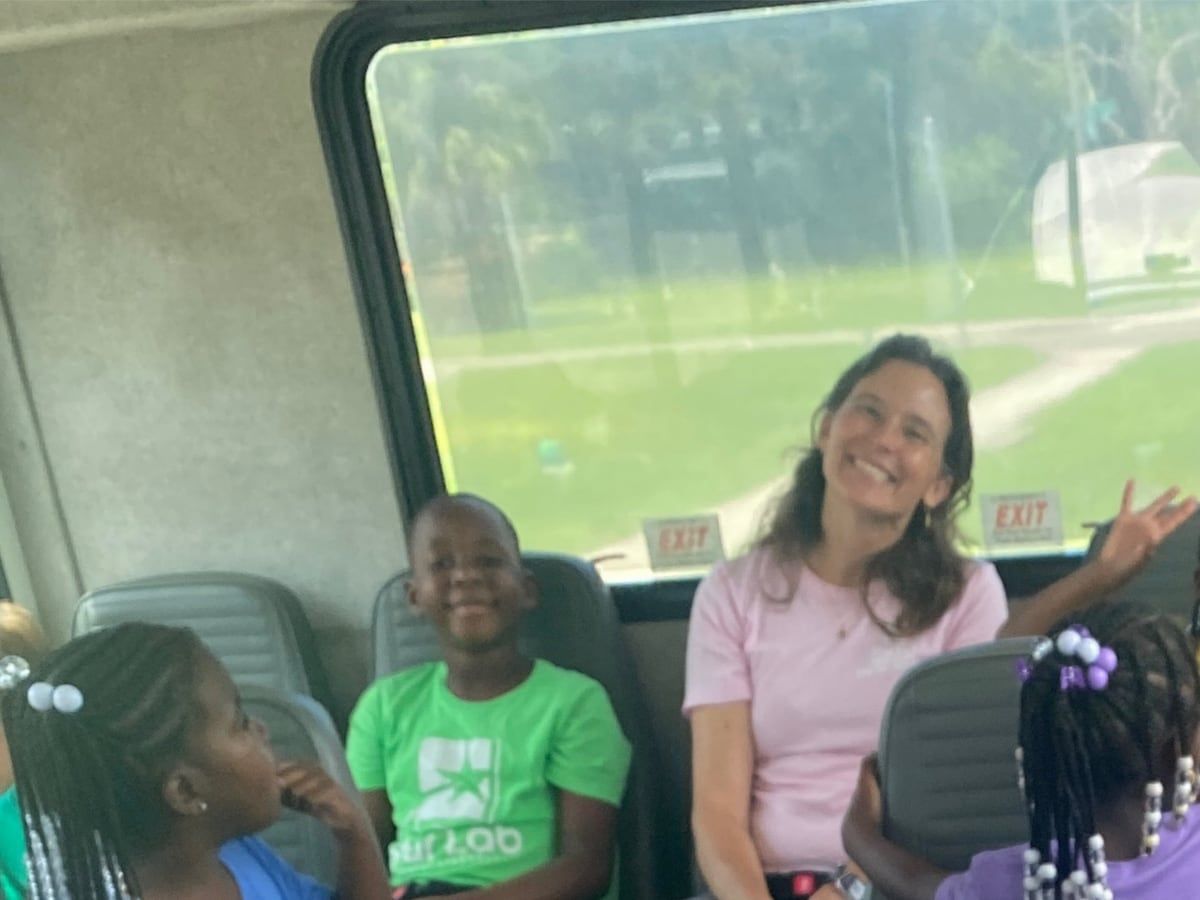 A smiling teacher and children seated on a bus; a boy in a green T-shirt grins at the camera.
