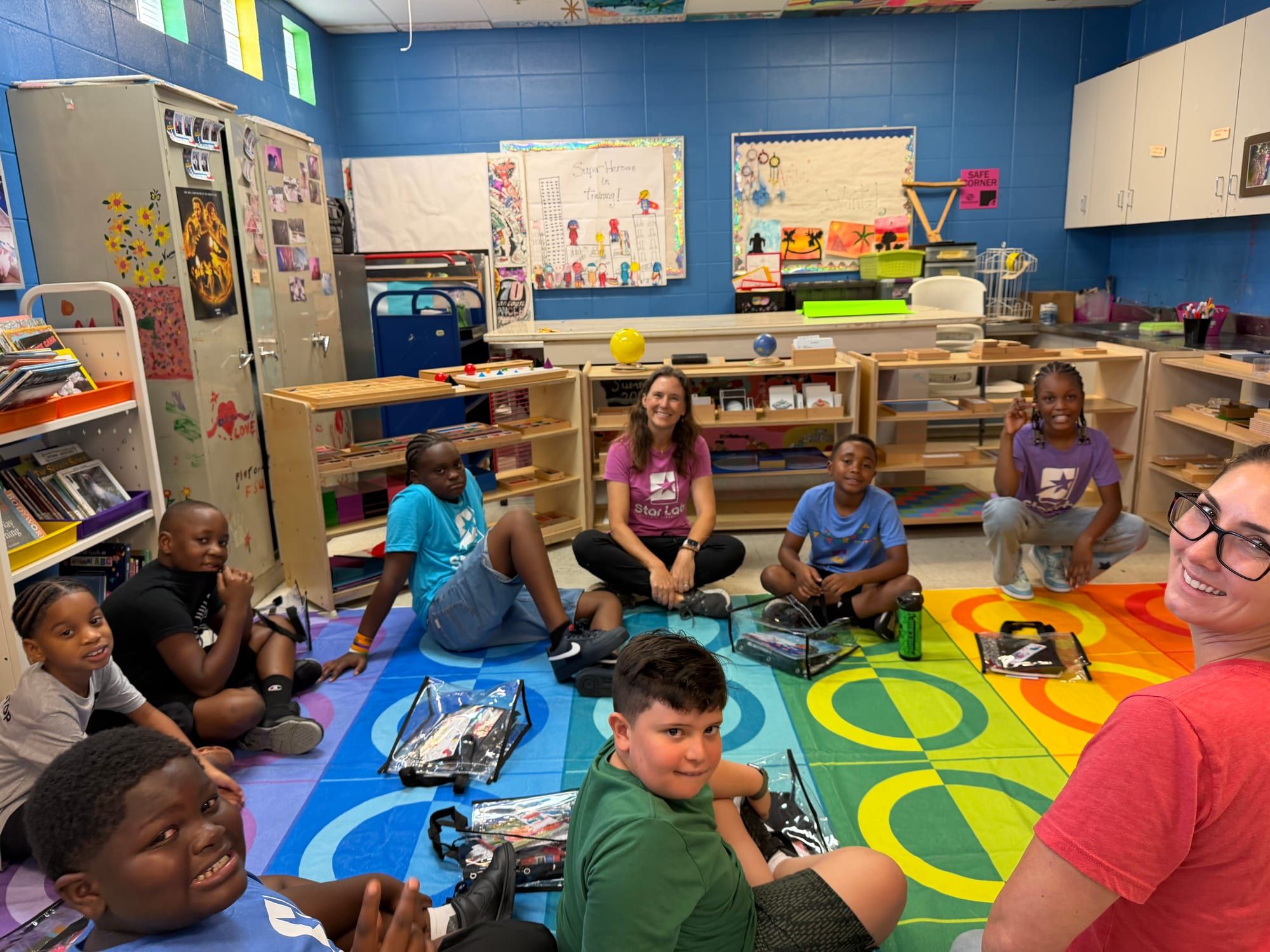 Children and teachers sit in a circle on a colourful rug in a blue-walled classroom with wooden Montessori shelves.
