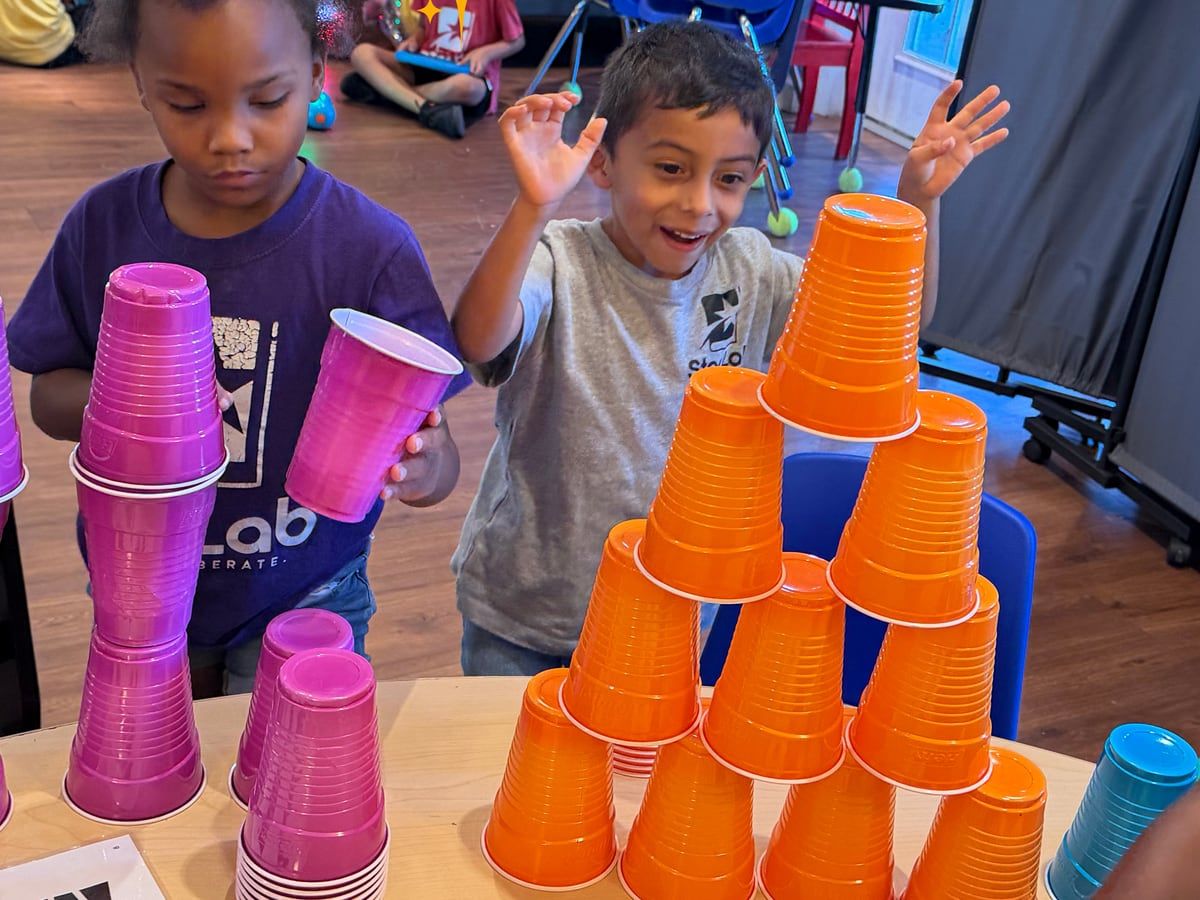 Children stacking pink and orange plastic cups into towers; a boy behind them cheers with both hands up.