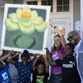 Alison Rini, in a purple Star Lab shirt, and children hold up a large painting of a yellow lotus flower outside the school at 2300 Central Ave.