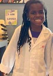 A smiling child wearing a white lab coat stands in a classroom.