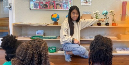 A young woman kneels in front of a low classroom shelf of Montessori materials and potted flowers, pointing at the shelf while seated children watch.