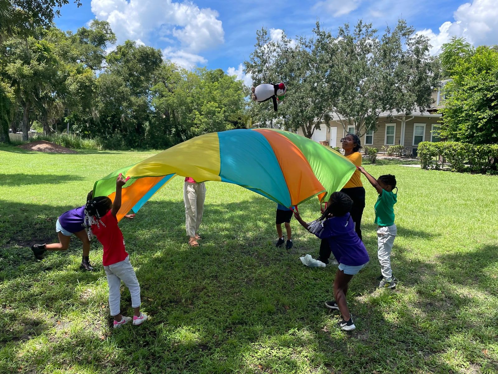 Children and two adults lift a rainbow-coloured play parachute on a sunny lawn, with a stuffed penguin toy tossed in the air above it and a house and trees behind.