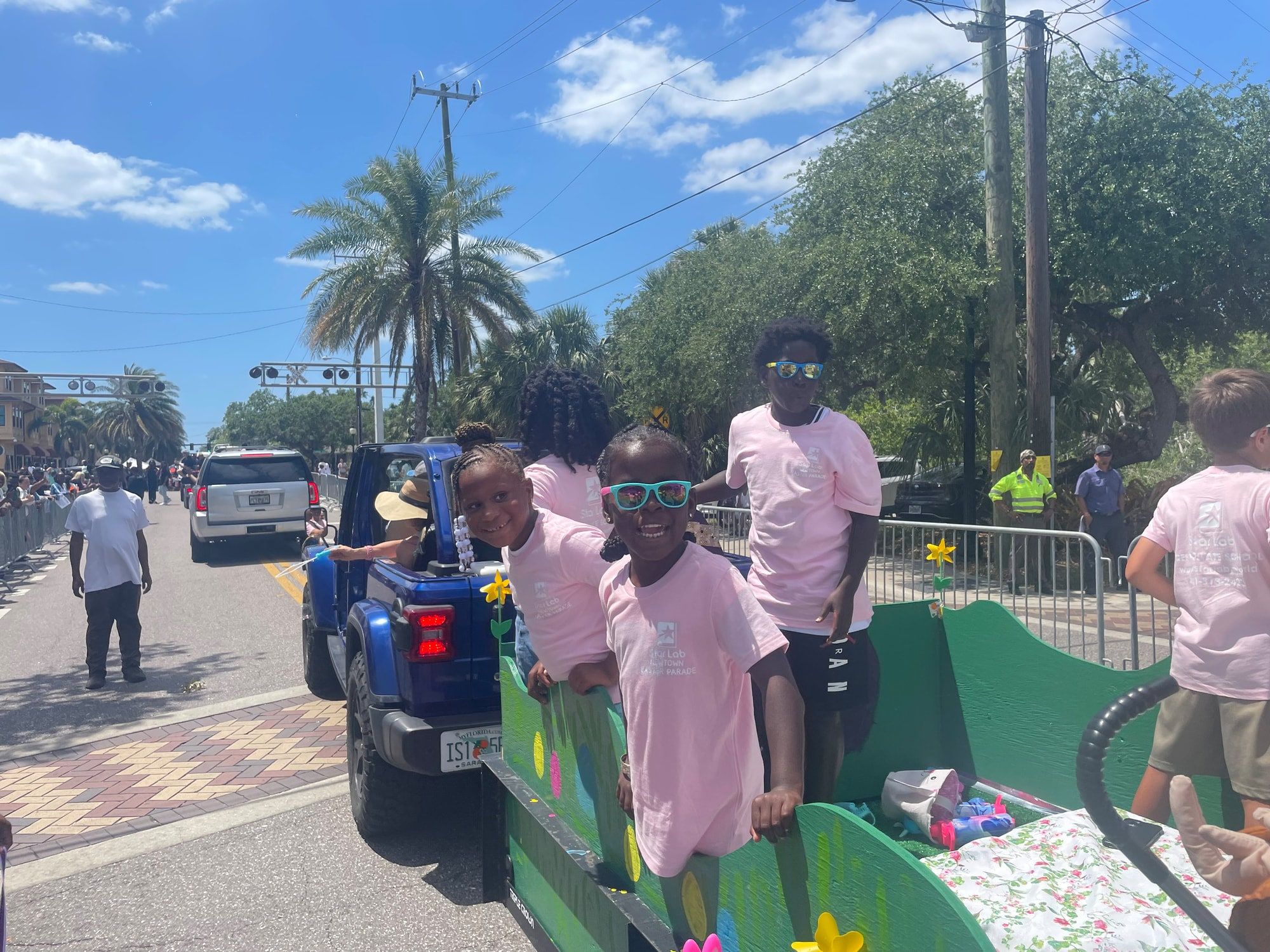 Children in pink Star Lab T-shirts ride on a decorated green float pulled by a blue Jeep along a street parade route lined with spectators and palm trees.