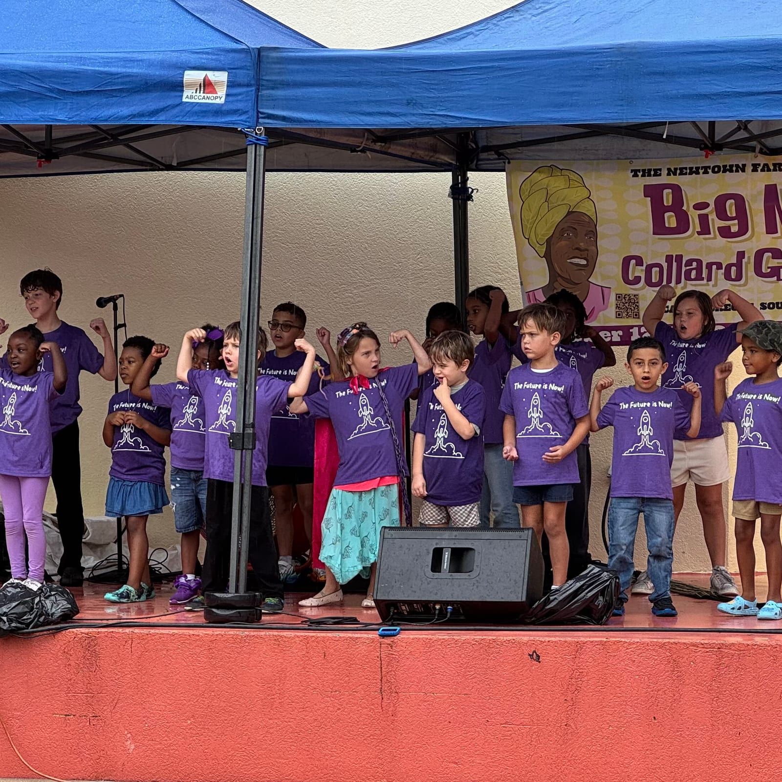 Children in purple Star Lab T-shirts perform on an outdoor stage under a blue canopy at the Big Mama Collard Greens Festival, flexing their arms.