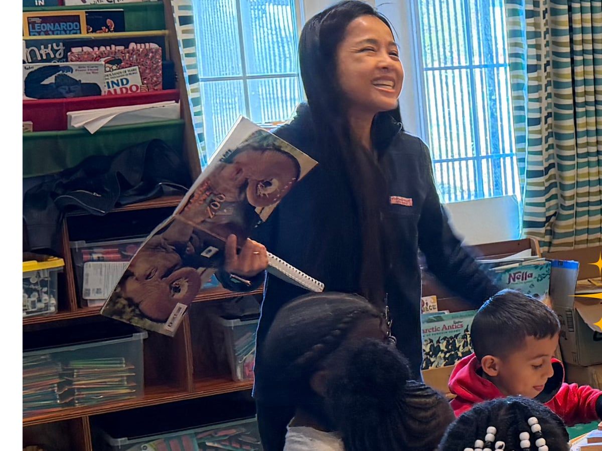 A smiling teacher holds up a picture book in front of the class bookshelves while children sit at a table.