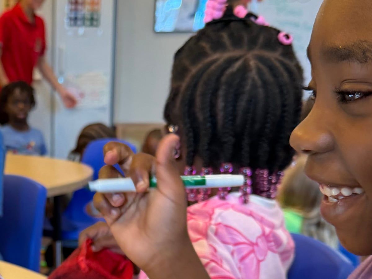 A smiling girl in class holding a green marker, with classmates working behind her.
