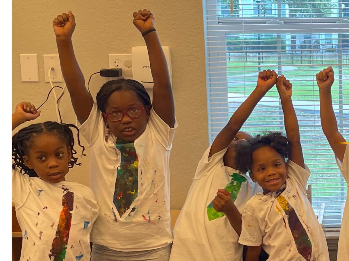 Children in white T-shirts with painted paper ties raise their fists in a classroom by a window.