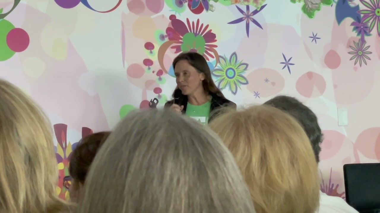 Alison Rini, in a green Star Lab T-shirt, speaks into a microphone in front of a colourful floral mural, seen over the heads of the audience.