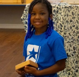 A smiling girl with blue-beaded braids, wearing a blue Star Lab T-shirt, sits holding a small wooden box.