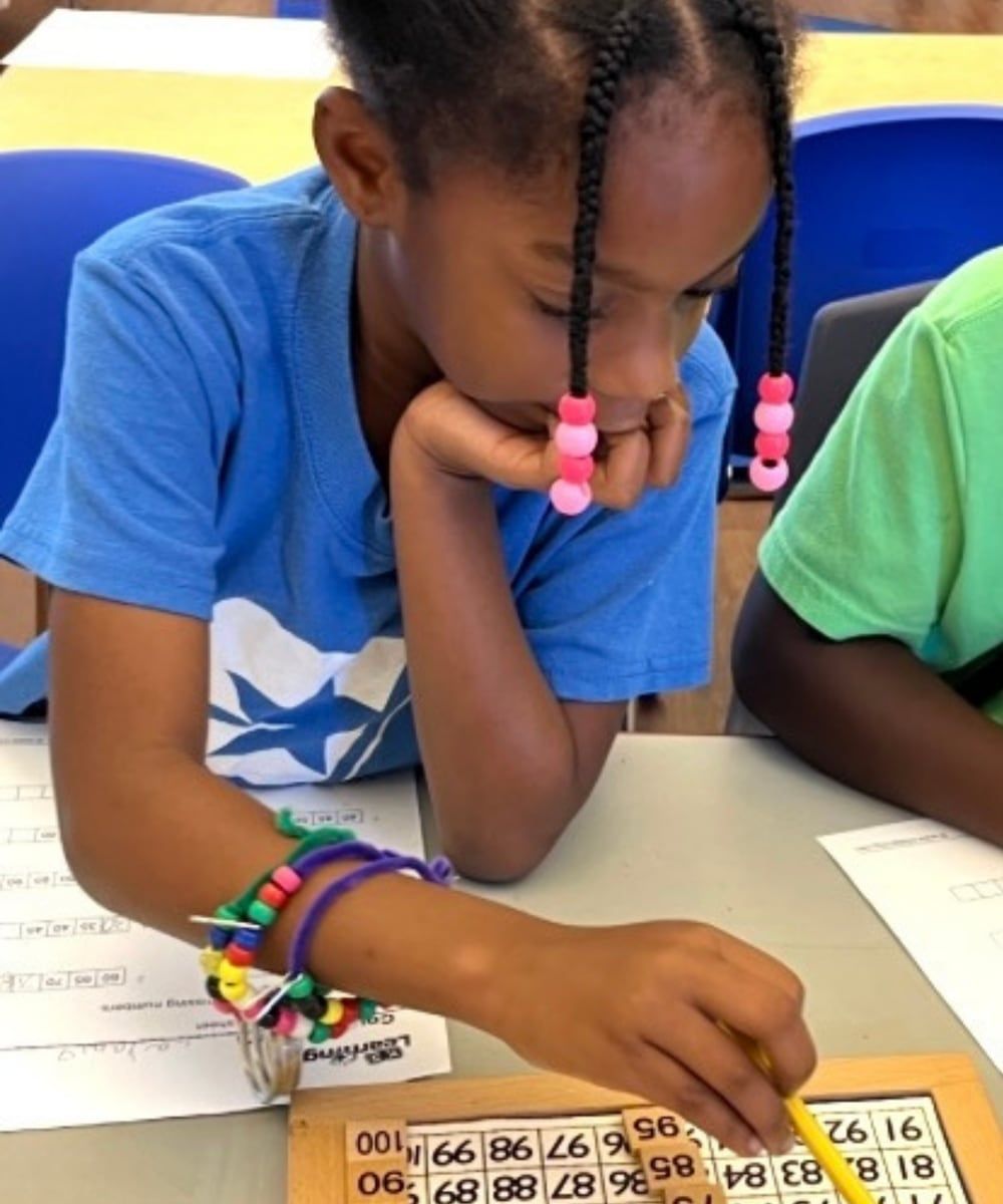 A girl in a blue Star Lab T-shirt rests her chin on her hand as she works through a number worksheet beside a wooden hundred-board puzzle.