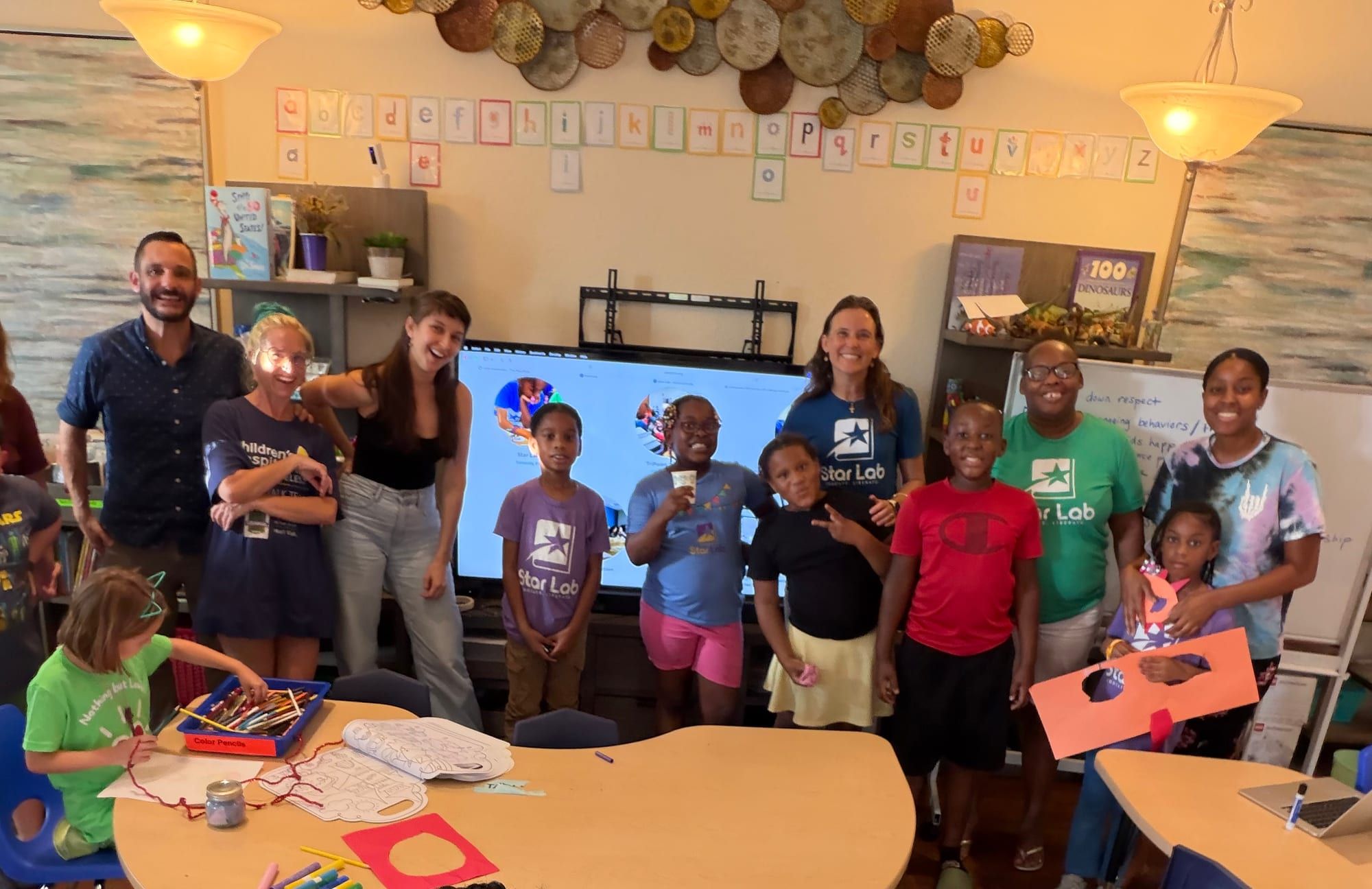 Group photo of teachers and children, several in Star Lab T-shirts, in a classroom with an alphabet banner and a large screen.